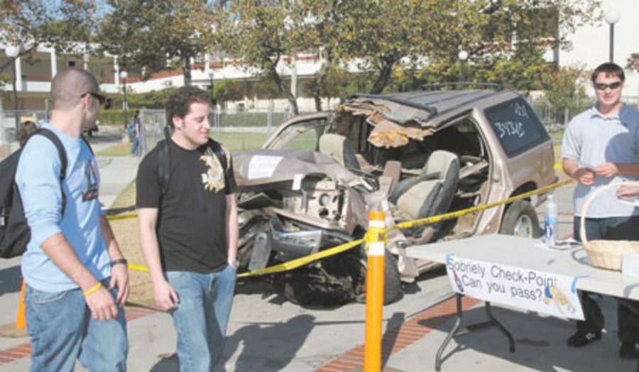 Crashed auto of 18 year old  MC student under the influence who died in the crash, with student on-lookers walking past, and a Sobriety Check-Point table to the right sponsored by the Student Health Center on campus.